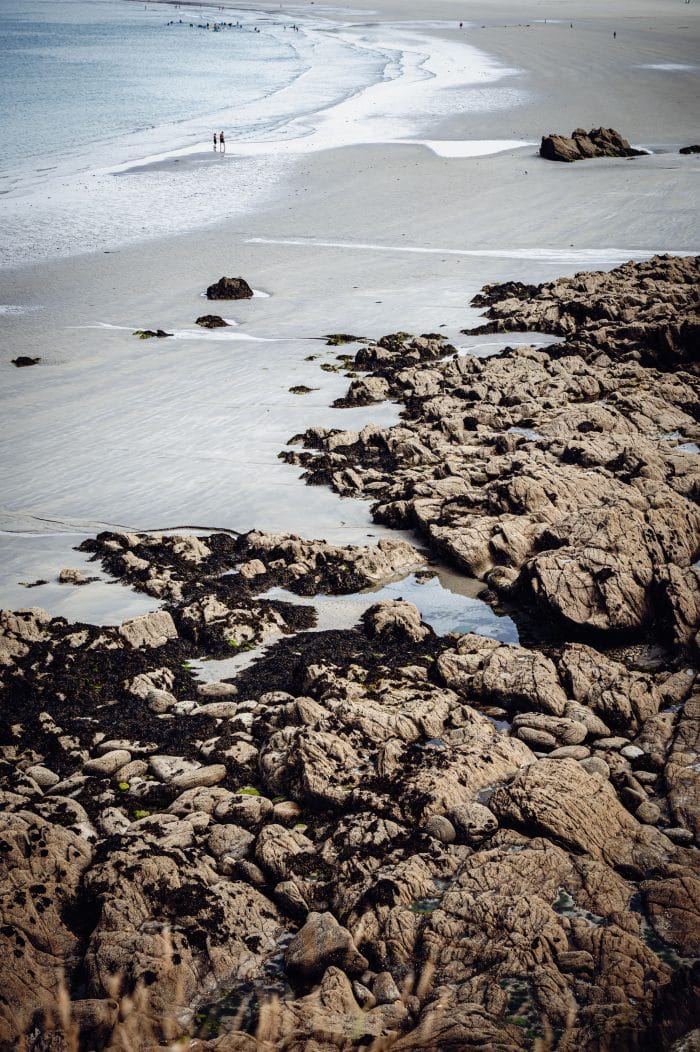 Raue Felsen im Vordergrund begrenzen den bretonischen Sandstrand an dem im Hintergrund zwei Menschen spazieren gehen.