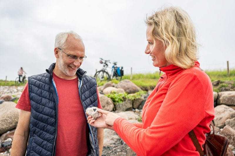 Eine Frau zeigt ihrem Mann einen Stein, in dem ein Herz zu sehen ist. In Hintergrund sieht man die Fahräder der beiden stehen.