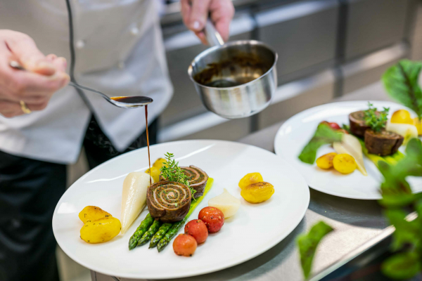 Herr Elbers, der Küchenchef, richtet einen Essensteller an. Gerade träufelt er Soße über das auf dem Teller angerichtete Fleisch. Aussagekräftige Fotos für Ihr Hotel-Unternehmen.