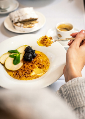 Die Hand eines Restaurantgastes hält einen Löffel mit Crème brûlée das er aus einem Dessert-Teller löffelt.