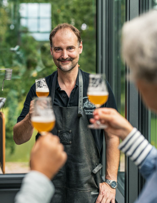 Der Brauerei-Inhaber, ein Mann in einer Brauerei-Schürze, prostet mit einem frisch gezapften Glas Bier zwei Gästen zu.