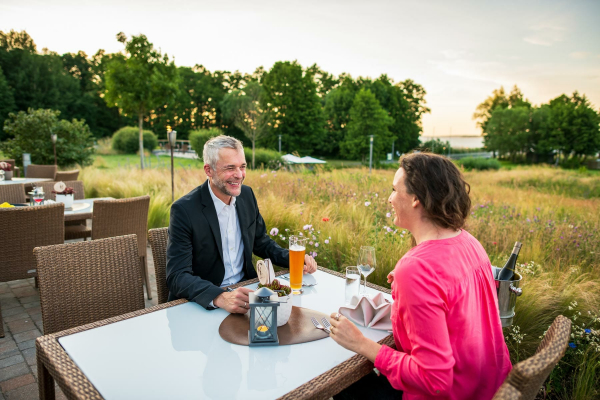 Aussagekräftige Fotos für Ihr Hotel-Unternehmen. Zwei Hotelgäste sitzen auf der Hotelterrasse an einem Tisch. Die Gäste freuen sich über den schönen Platz. Hinter der Terrasse sieht man in die schöne Landschaft die das Hotel umgibt.
