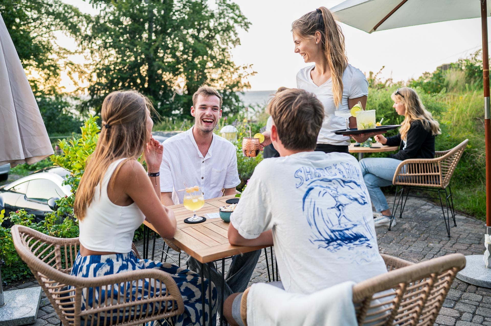 ein Tisch mit gut gelaunten jungen Leuten wird von einer Hotelmitarbeiterin mit Cocktails bedient