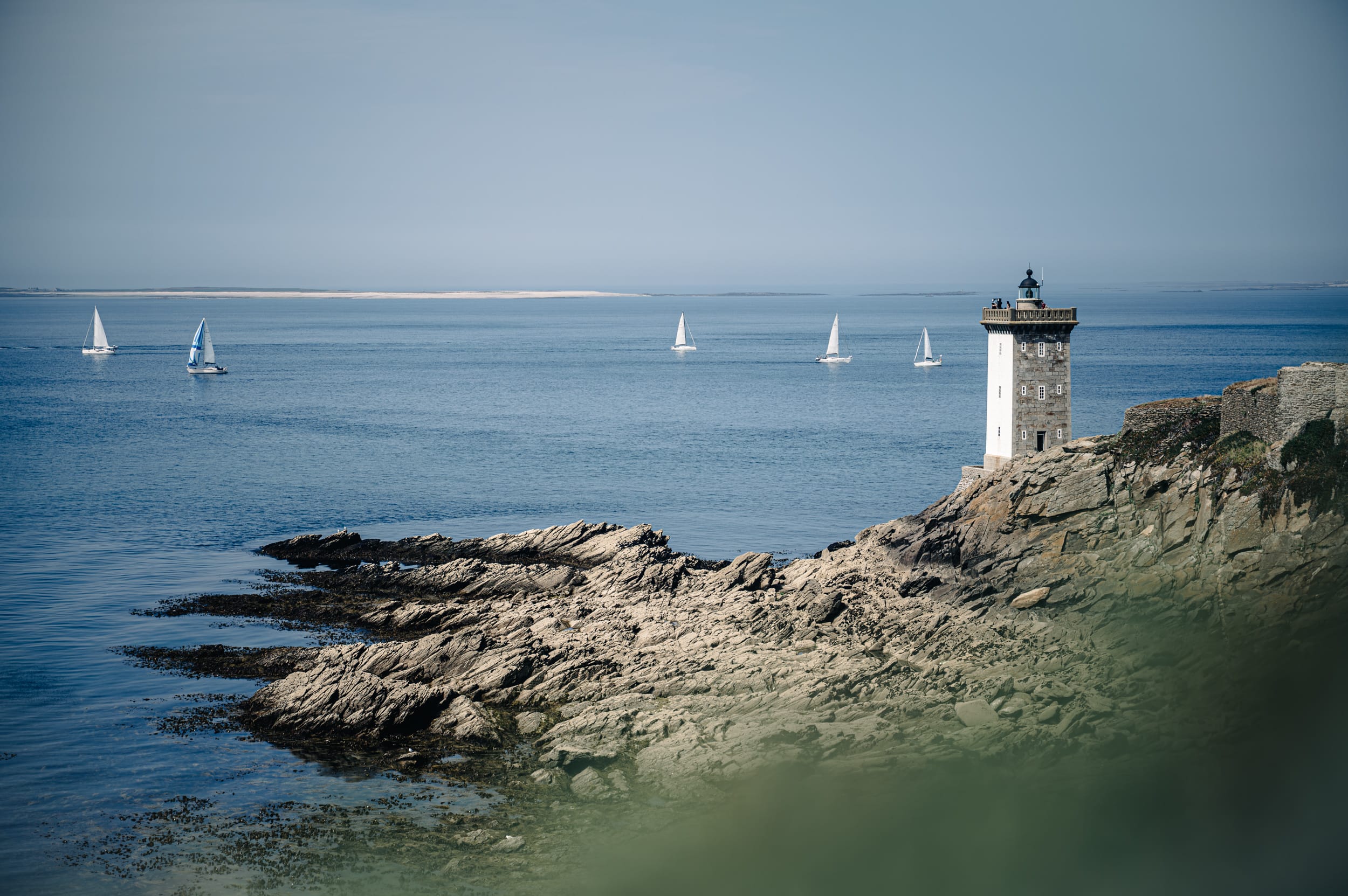 Leuchtturm an schroffer bretonischer Küste mit Segelbooten im Hintergrund