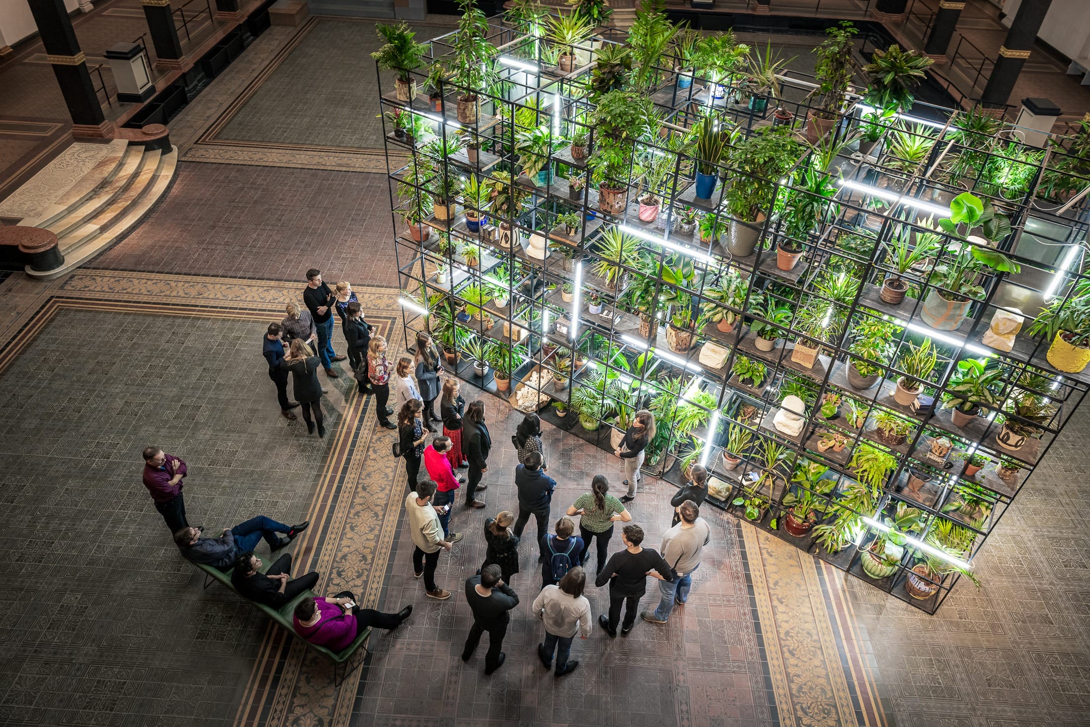 Ein riesiges Regal mit Topfpflanzen steht im Martin Gropius Bau in Berlin. Eine Gruppe von Menschen lassen sich von eine Museumspädagogin die Installation erklären.