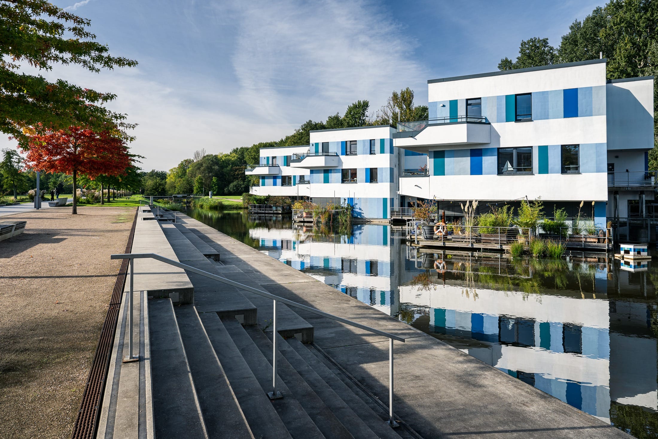 moderne weißblaue schwimmende Häuser auf einer Wasserfläche in Wilhelmsburg mit dem Wasser vorgelagerten Sitzstufen und Parkgelände. Foto vom Architekturfotografen